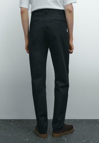 Pantaloni neri realizzati in tessuto liscio con un design a gamba dritta, dotati di due tasche posteriori e indossati con scarpe marroni.