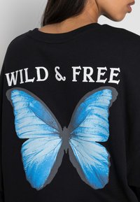 Felpa nera con una grande grafica di una farfalla blu sul retro, con il testo "WILD & FREE" in bianco sopra il design della farfalla.