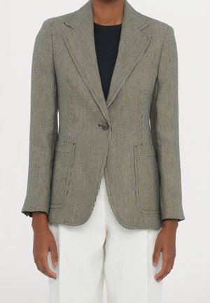 Persoon draagt een zwart-wit geruite blazer, eenmaal dichtgeknoopt, met zakken aan de voorkant, over een zwart topje en witte broek.