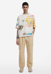 Majica s okruglim izrezom i kratkim rukavima s apstraktnim akvarelnim uzorcima u narančastoj, žutoj i zelenoj boji, u kombinaciji s bež cargo hlačama i smeđim tenisicama.