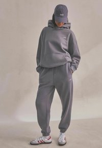 Szara bluza z kapturem i dopasowane dresy wykonane z miękkiego materiału, z przednią kieszenią i akcentem z logo. Szara czapka dopełnia outfit.