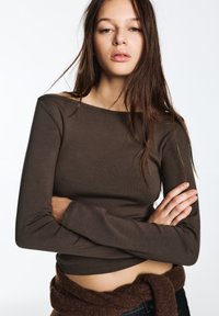 Top marrone a maniche lunghe, aderente, con collo ampio e trama a coste; valorizzato da una cintura in maglia marrone in vita.