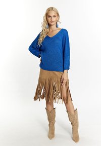 Mėlynas megztinis su V formos iškirpte, derinamas su smėlinio spalvos sijonu su frengais. Bežiniai kaubojiški batai pabrėžia aprangą, demonstruodami vakarietišką stilių.