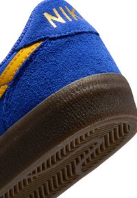 Kék velúr sportcipő sárga részletekkel, texturált barna gumitalppal és arany színű "NIKE" logóval a saroknál.