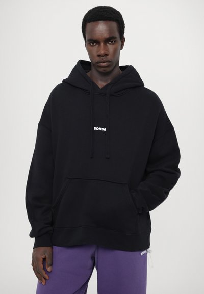 Črn oversize kapucar iz bombaža, ki ima kenguru žep in majhen bel logotip na sredini. V kompletu s vijoličnimi trenirkami.