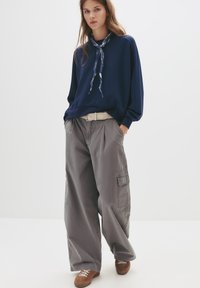 Felpa blu, pantaloni cargo grigi con tasche laterali, cintura beige e scarpe marroni, con una sciarpa a motivi blu annodata al collo.