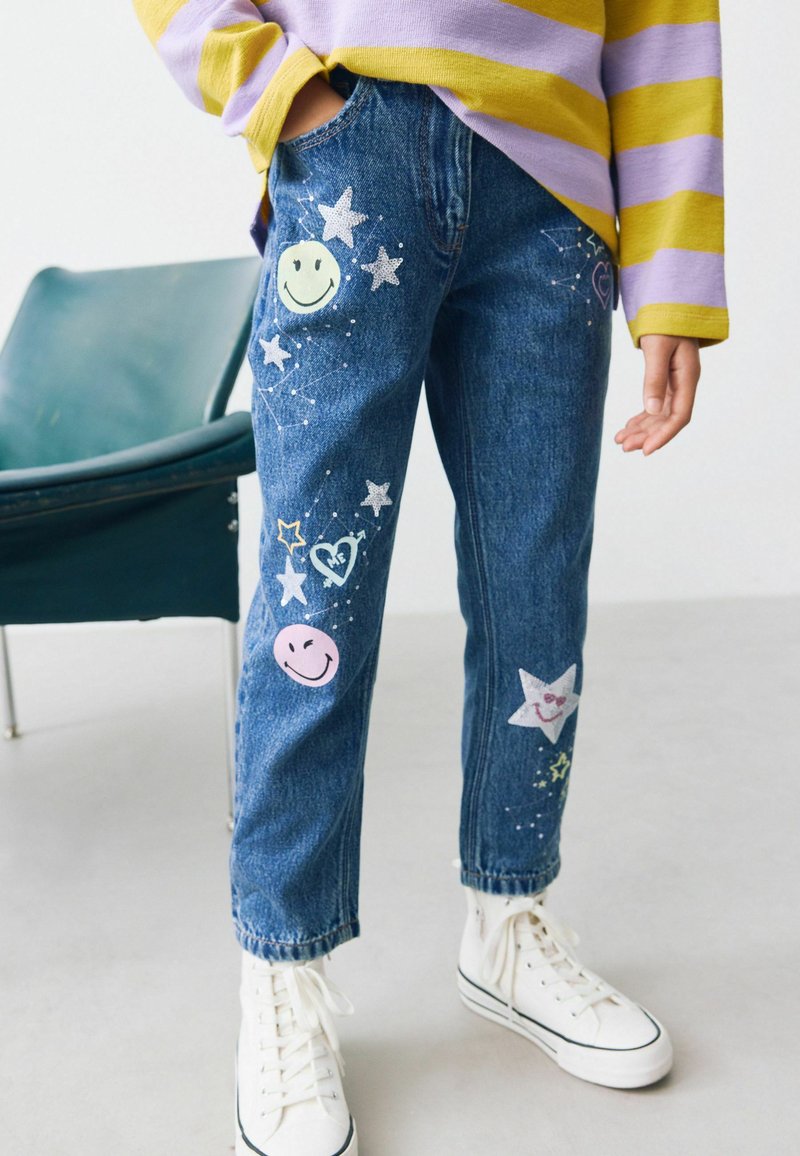 Denim jeans met kleurrijke smileygezichten, sterren en hartjes die op de voorkant zijn aangebracht, met een relaxte fit en enkel lengte, gecombineerd met witte sneakers.