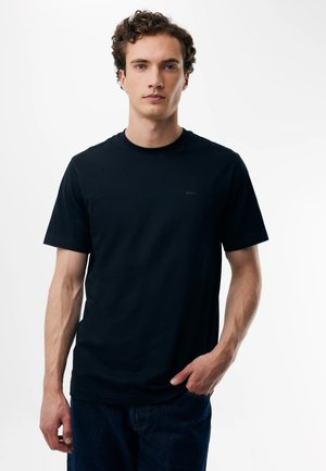 Giovane uomo con capelli ricci che indossa una t-shirt nera a maniche corte e jeans scuri su uno sfondo bianco semplice.