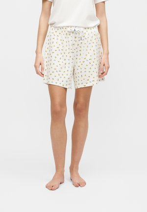 Γυναίκα που φοράει λευκά σορτς πυτζάμα με μικρά κίτρινα τυπώματα λεμονιού και λευκό κορδόνι δεμένο στη μέση, συνδυασμένα με απλό λευκό μπλουζάκι.