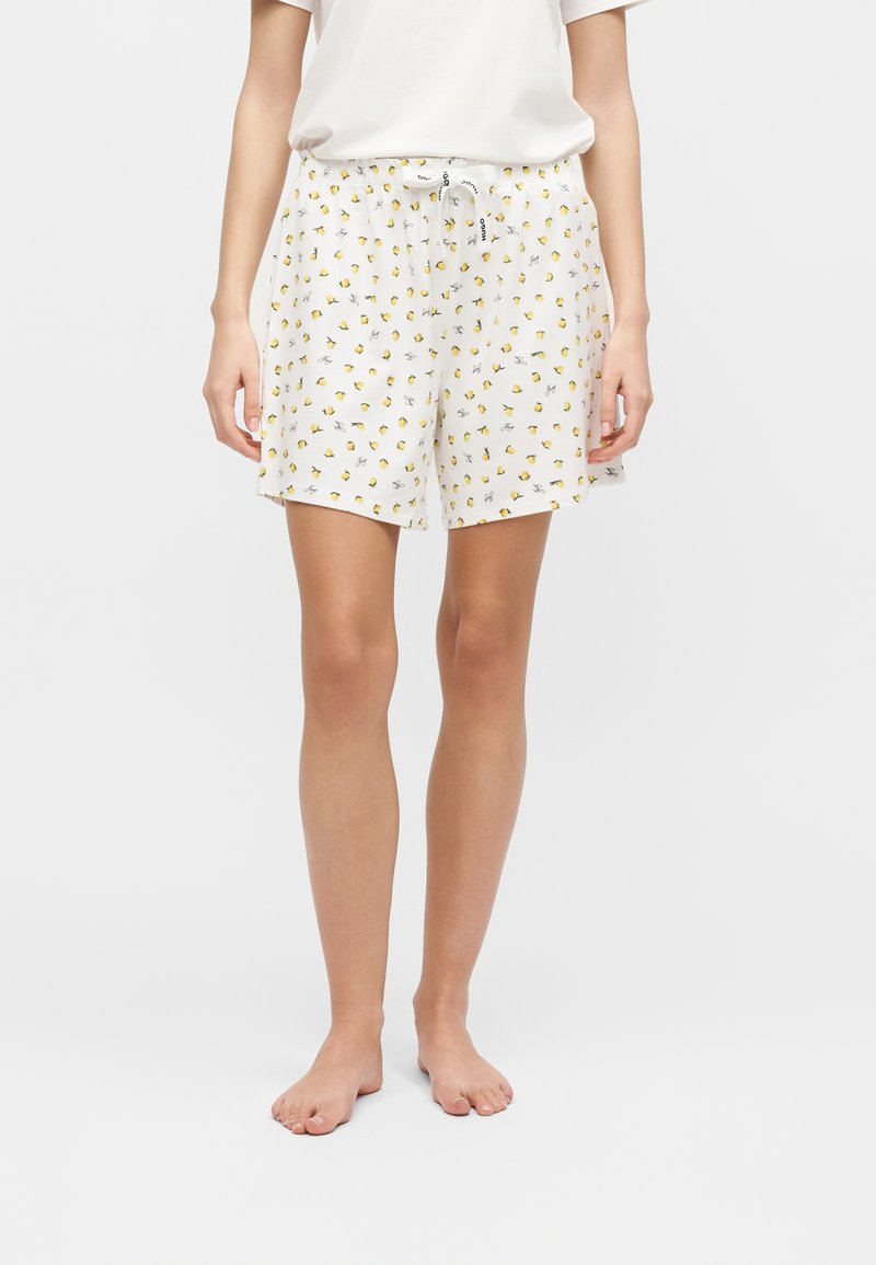 Γυναίκα που φοράει λευκά σορτς πυτζάμα με μικρά κίτρινα τυπώματα λεμονιού και λευκό κορδόνι δεμένο στη μέση, συνδυασμένα με απλό λευκό μπλουζάκι.