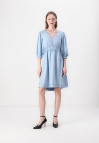 Rochie din denim de culoare bleu deschis, cu decolteu adânc în formă de V, mâneci bufante și talie strânsă. O pereche de pantofi cu toc înalt negri completează look-ul.