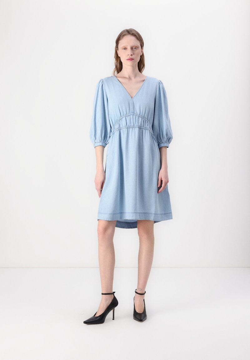 Rochie din denim de culoare bleu deschis, cu decolteu adânc în formă de V, mâneci bufante și talie strânsă. O pereche de pantofi cu toc înalt negri completează look-ul.