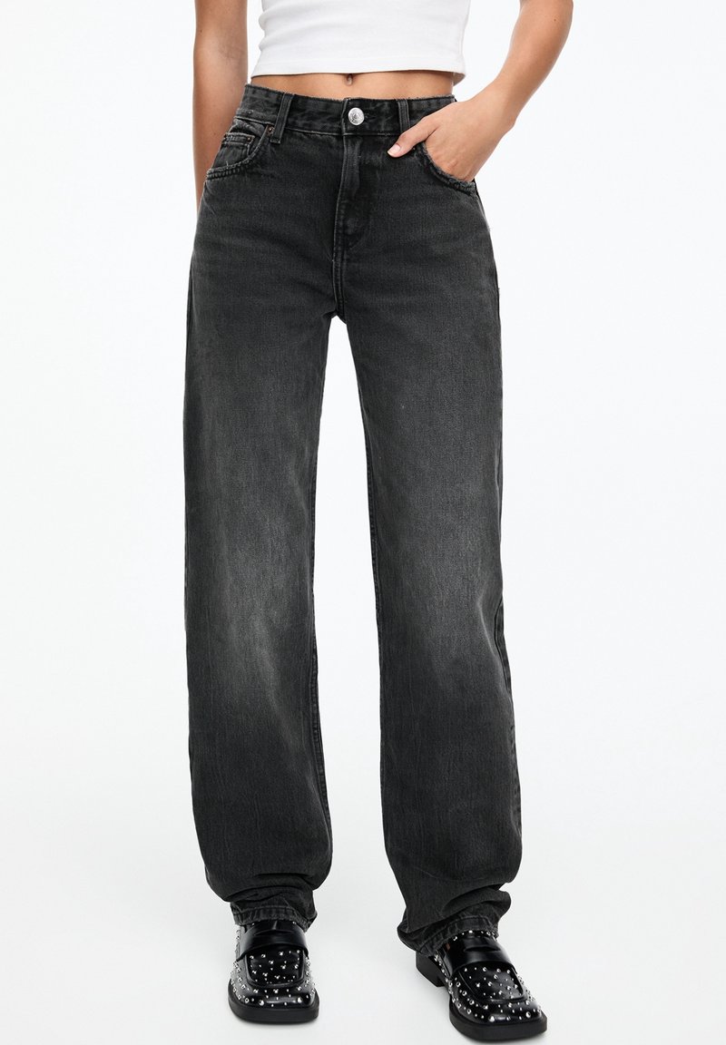 Femme portant un jean noir taille haute, ample, un top court blanc et des mocassins noirs cloutés, une main dans la poche.