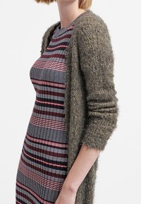 Grön, fluffig cardigan över en åtsittande, randig klänning i rött, svart och vitt. Texturerat stickat tyg med varierande linjemönster.