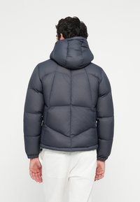 Giacca a vento grigia scura con cappuccio, caratterizzata da un design trapuntato, polsini a coste e una vestibilità leggermente accorciata. Tessuto liscio e isolante.