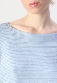 Lyseblå topp med båthals, med teksturert, ribbestrikket mønster og mykt stoff, som viser skuldre og nakke.