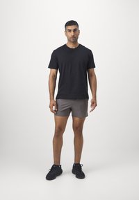 Černé tričko s krátkým rukávem v kombinaci se šedými šortkami. Model má na sobě černé sportovní boty. Jednoduché pozadí zvýrazňuje oblečení.