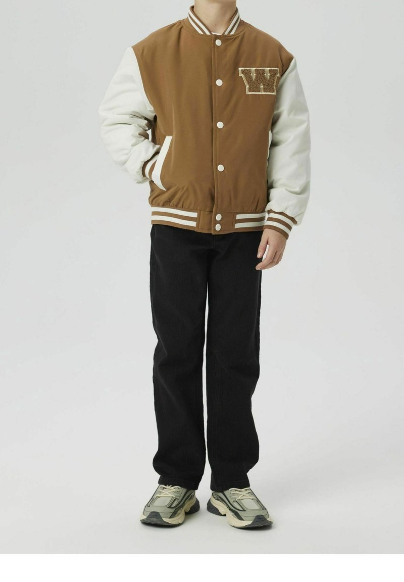 Personne portant une veste universitaire marron avec des manches blanches, un pantalon noir et des baskets grises, debout avec une main dans la poche sur un fond blanc.