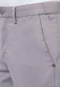 Jasnoszare spodnie wykonane z teksturowanej tkaniny mają płaski przód, subtelny zagięcie oraz ozdobny guzik z logo marki.