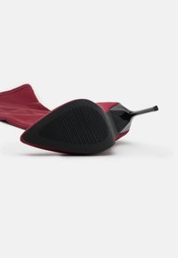 Stivale rosso al ginocchio con tacco a stiletto, dalla punta affusolata e sopra liscia. La suola è nera con texture a righe, caratterizzata da un tacco sottile e affusolato.
