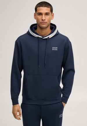 Mann trägt einen marineblauen Hoodie und passende Jogginghose mit weißen "Joop! Jeans" Logos auf Brust und Oberschenkel, steht vor einem einfarbigen Hintergrund.