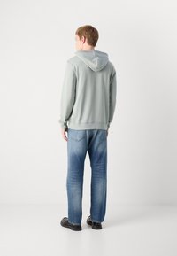 Jonge man die met zijn rug naar de camera staat, gekleed in een lichtgrijze hoodie, blauwe versleten jeans en zwarte schoenen, tegen een plain witte achtergrond.