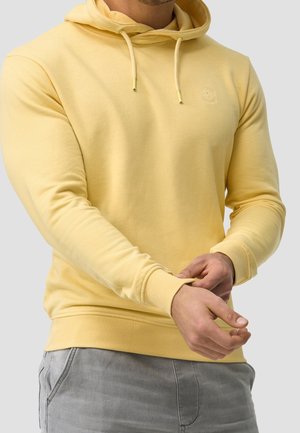 Homme portant un sweat à capuche jaune clair ajustant le poignet avec les deux mains, vêtu d'un pantalon gris clair, sur un fond uni.