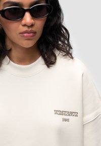 Sweatshirt crema con scollatura a costine, tessuto morbido e logo ricamato marrone sul petto. Indossato con occhiali da sole ovali scuri.