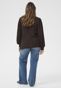 Kvinde med langt brunt hår iført en mørk langærmet bluse og blå jeans, stående med ryggen til foran en ensfarvet hvid baggrund.