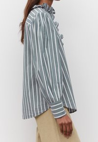 Camicia a righe blu chiaro e bianca con colletto a balze e maniche lunghe a sbuffo. Realizzata in tessuto morbido, presenta polsini con bottoni e un fit svasato.