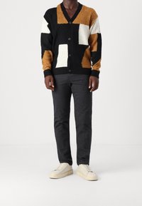Kardigan w blokach kolorów - czarny, biały i złoty, o teksturze warkoczowego splotu, zapinany na guziki, o luźnym kroju. Połączony z ciemnymi spodniami i jasnymi butami.