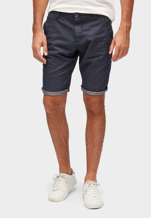 Homme portant un short bleu marine retroussé, des baskets blanches et un t-shirt blanc, se tenant devant un fond uni clair.