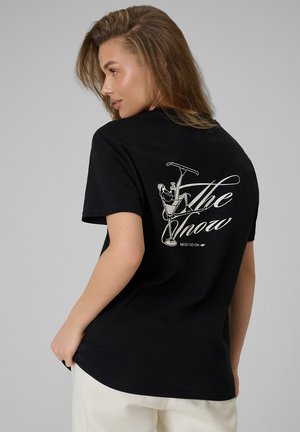 Kobieta w czarnej koszulce z białą grafiką przedstawiającą osobę odpoczywającą w kieliszku do martini oraz tekstem "The Show must go on."