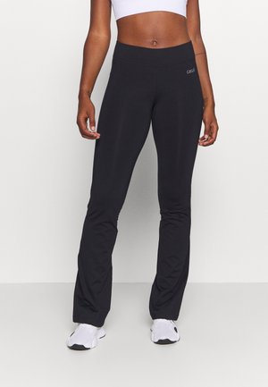 Vrouw die strakke zwarte uitlopende sportbroek en witte sneakers draagt, staand tegen een effen achtergrond.