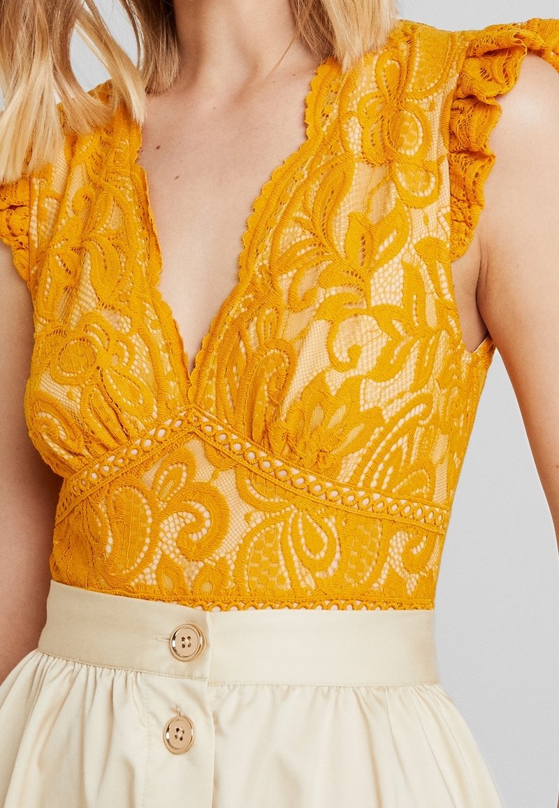 Gul spetsblus med blommönster och volangärmar, med en djup V-ringning, tillsammans med en krämfärgad knäppt kjol.