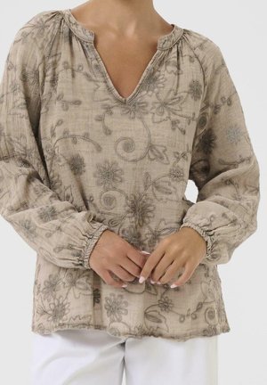 Blouse beige à manches longues avec broderie florale et poignets froncés portée avec un pantalon blanc, mains croisées à la taille.