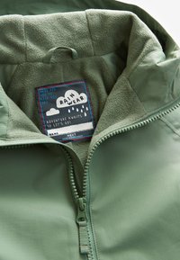 Grøn regnjakke med fleecefor, delvist åbnet lynlås, der viser label med regnsky og regndråber grafik samt tekst om at holde sig tør.