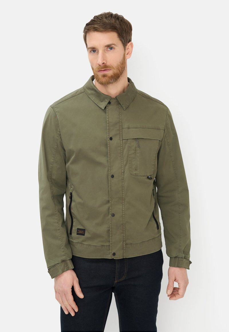 Man met baard, gekleed in een olijfgroene jas met voorzakken en een zwarte broek, staand tegen een effen lichte achtergrond.