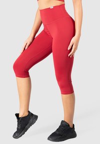 Leggings capri rouges fabriqués en tissu extensible avec une taille haute. Présentent un design sans coutures et des jambes fuselées. Des baskets noires complètent la tenue.