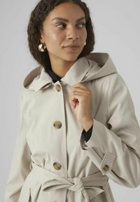 Trench-coat imperméable beige avec une capuche, boutons dorés, ceinture à la taille et poignets retroussés. Texture lisse et silhouette ajustée.