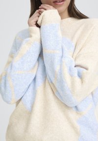 Kerma- ja vaaleansininen pehmeä, pörröinen neule, jossa geometrisia kuvioita ja resorit hihansuissa. Rennon istuvuuden omaava pyöreä kaula-aukko.