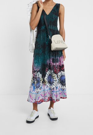 Mujer con vestido largo sin mangas de flores en tonos teal y púrpura, zapatillas blancas, sosteniendo una chaqueta blanca y un bolso bandolera color crema con la palabra "Love".