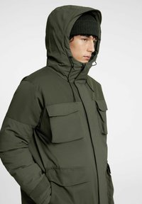 Olivgrön vadderad jacka med huva, med två stora bröstfickor och dragkedja i mitten. Texturerat tyg med justerbara dragskor.