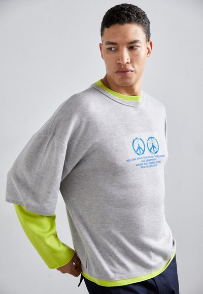 Sivina oversized pulover z modrim grafičnim besedilom in dodatki. Dolgi neonsko zeleni rokavi znotraj, zaokrožen izrez in sproščen kroj.