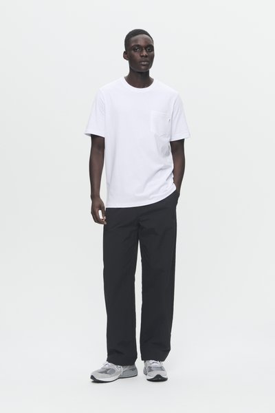 Balts kokvilnas T-krekls ar krūšu kabatu, apvienots ar brīviem melniem biksēm un pelēkiem sporta apaviem ar melniem akcentiem.