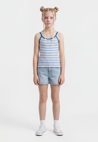 Girl with blonde hair in two buns and braids, wearing a white and blue striped tank top, light denim shorts, white socks, and white sneakers.