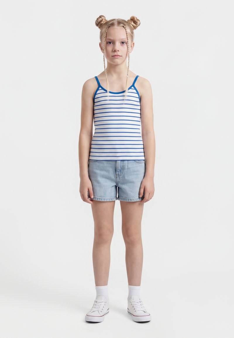 Girl with blonde hair in two buns and braids, wearing a white and blue striped tank top, light denim shorts, white socks, and white sneakers.