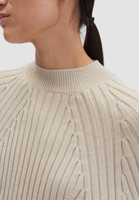 Primo piano di una persona con capelli scuri raccolti, che indossa un maglione a costine beige con un alto collo rotondo.