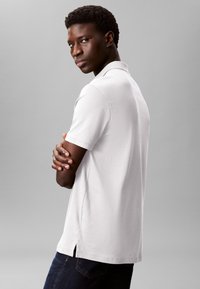 White short-sleeve polo shirt with a classic collar, side slits, worn with dark denim jeans. Model is viewed from the side against a gray background.