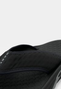 Κοντινό πλάνο μιας μαύρης σαγιονάρας με επενδεδυμένο ύφασμα στη ζώνη και υφασμένο πέλμα, σε απλό ανοιχτό φόντο.